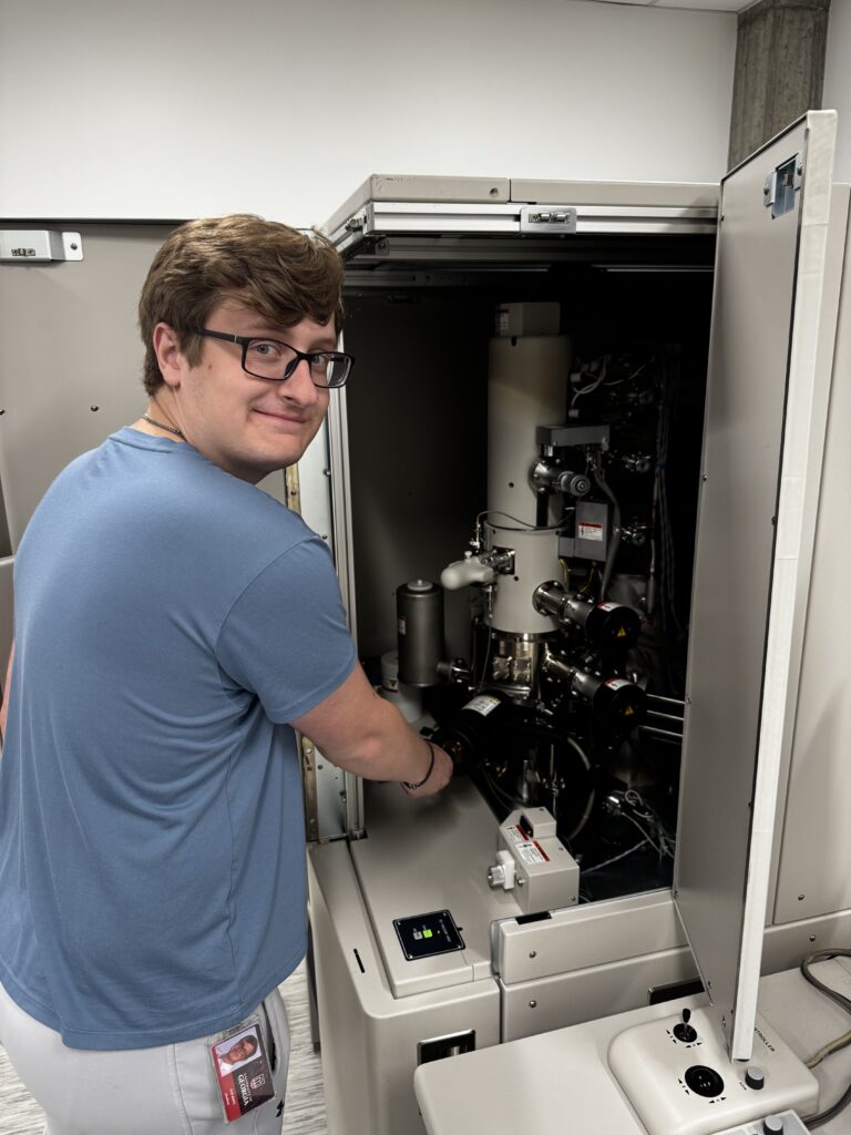 A person in a blue shirt operates a scientific instrument, possibly an electron microscope, in a laboratory setting.
